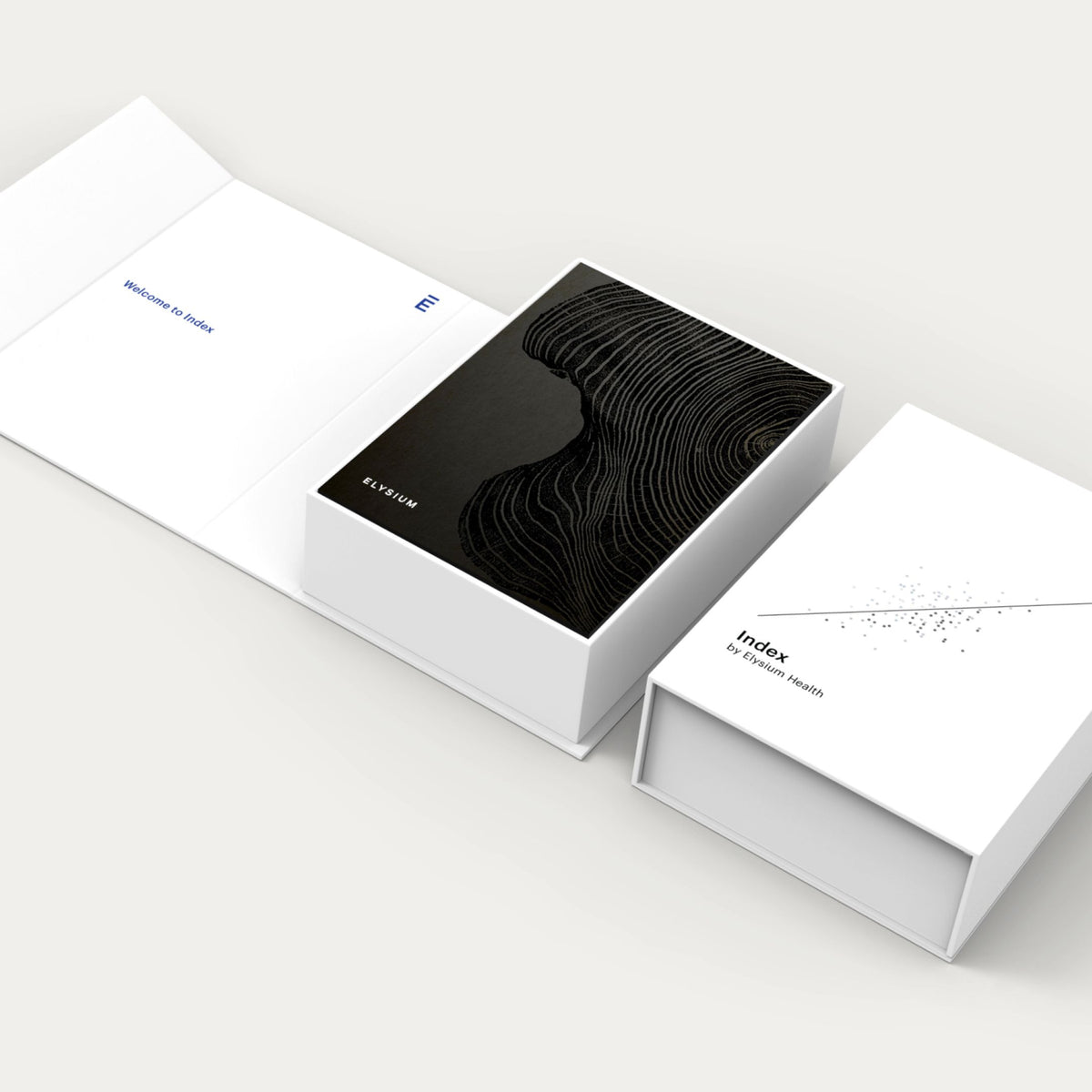 Three white product boxes arranged on a light gray studio background. The center box is open and reveals a black insert with a dark topographic line pattern; the right box is closed with the word 'Index' and small text beneath; the left box is open with blue text and a small blue mark.
