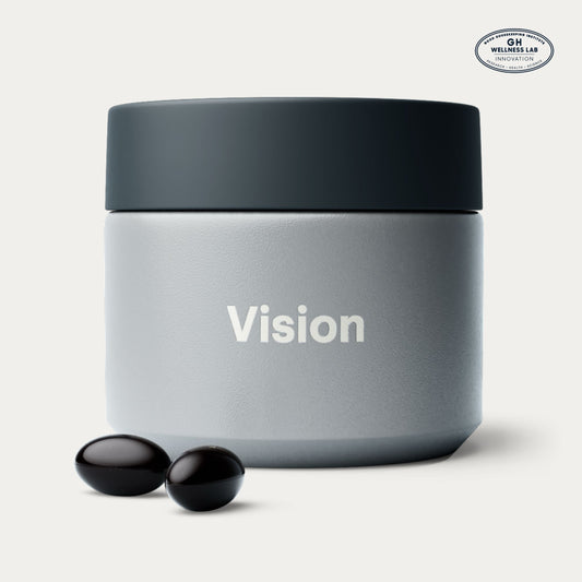 A matte gray cosmetic jar with a dark charcoal lid, featuring the word 'Vision' printed in white on the front. Two small black softgels are placed in front of the jar on a clean, light off-white background.